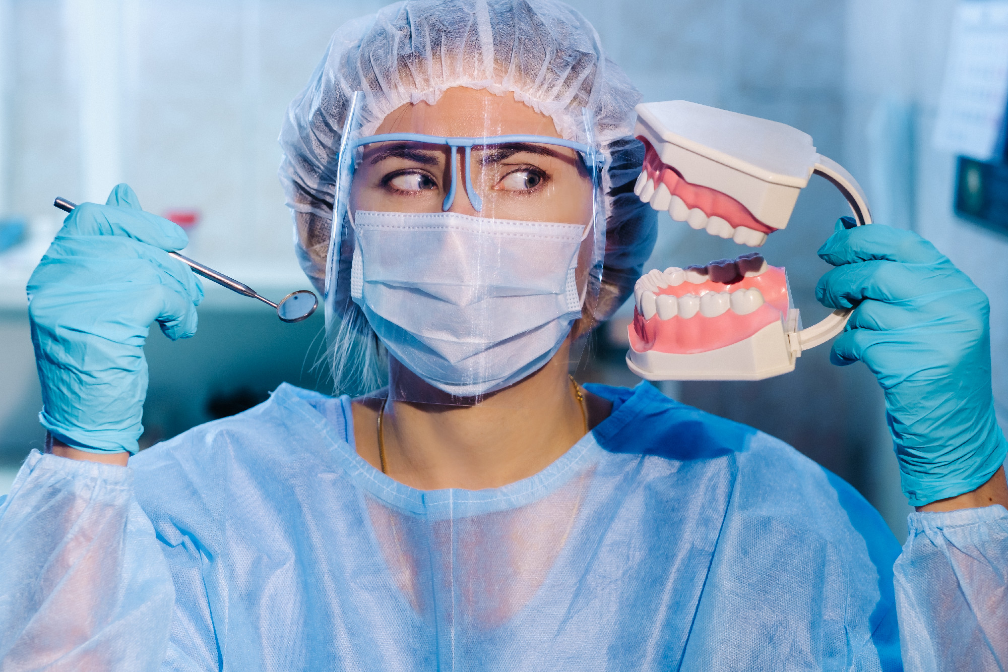 A dental doctor wearing blue gloves and a mask holds a dental model of the upper and lower jaws and a dental mirror