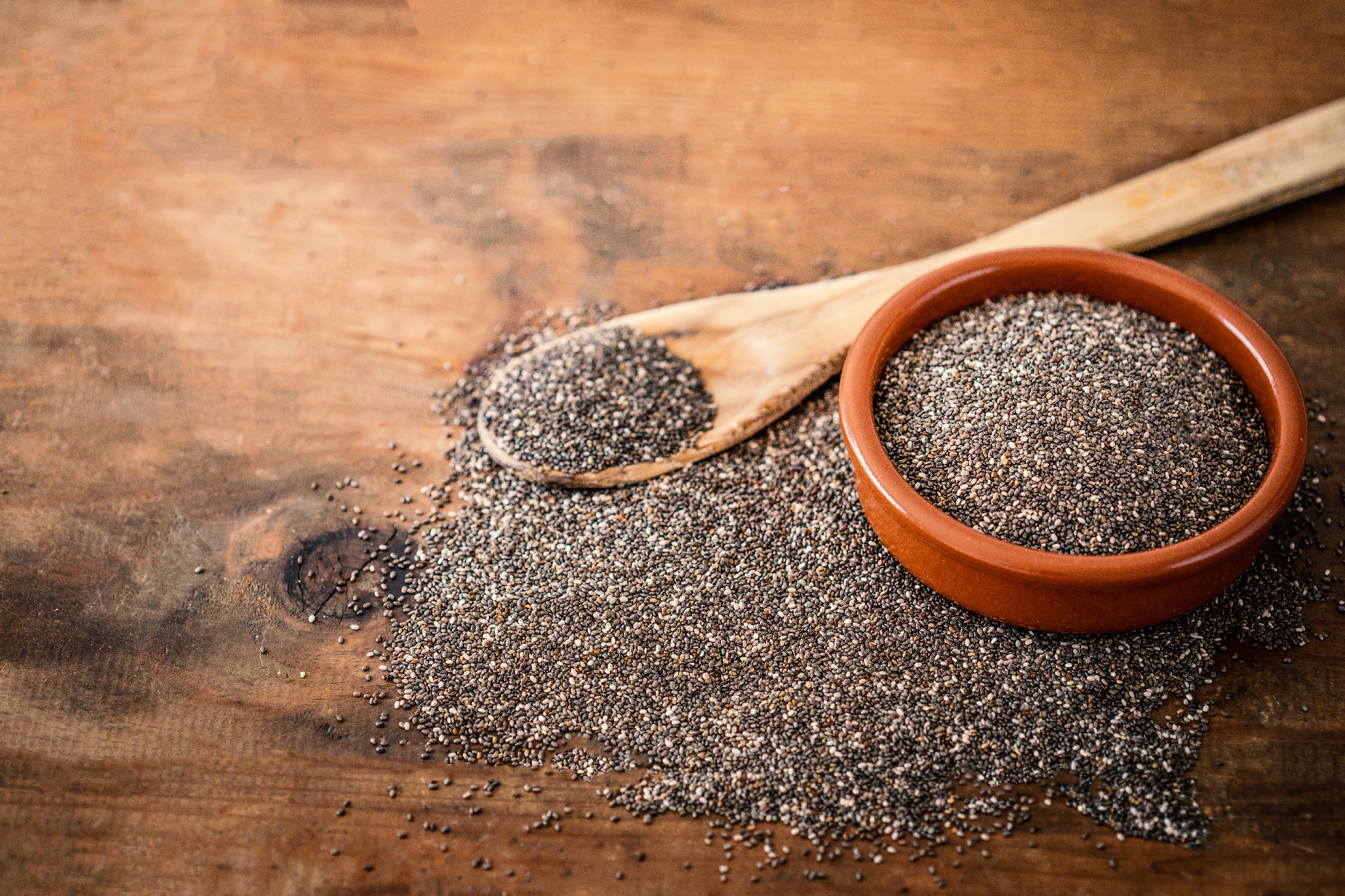 Chia seeds in a clay bowl and wooden spoon on wood background.