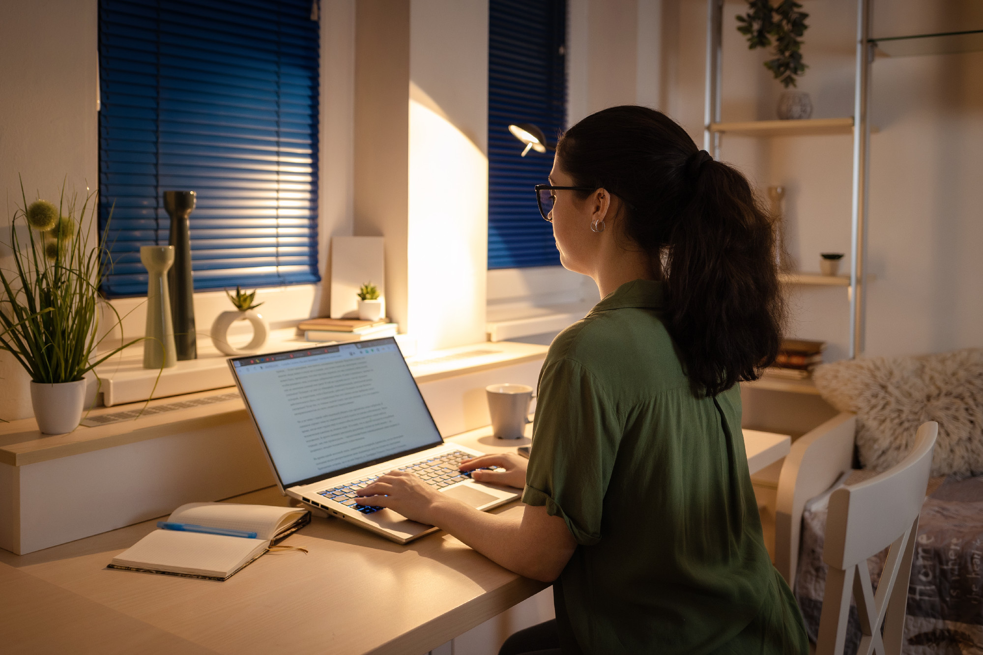 Online education. Back view of student young Caucasian woman working at laptop. Evening in home office with electric light. Concept of freelance and remote work
