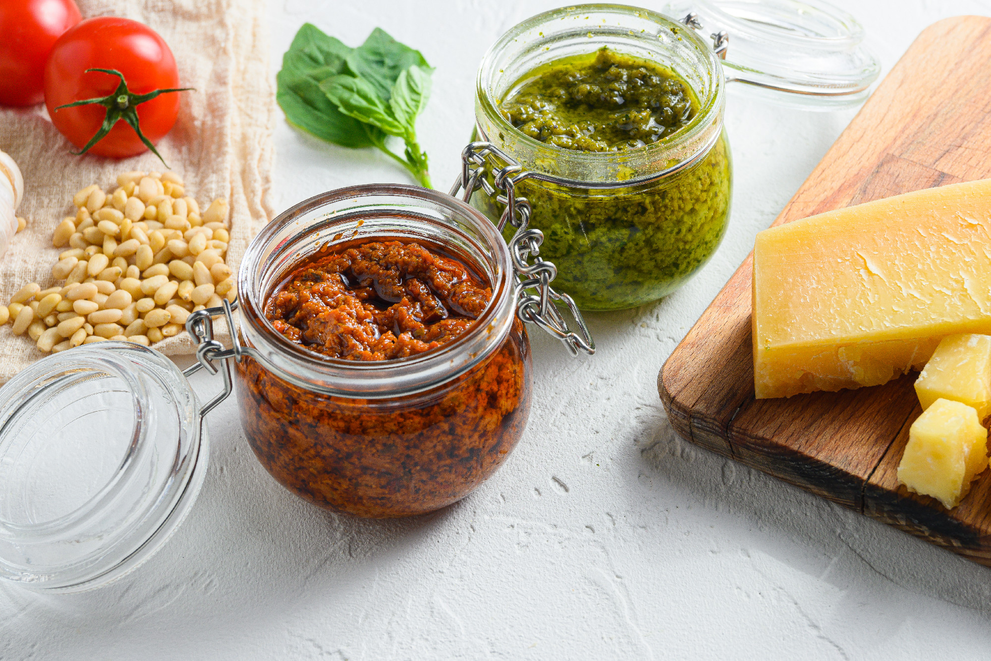 Glass jars with red and green pesto and cooking ingredients Parmesan cheese, basil leaves, pine nuts, white stone background, close up side view