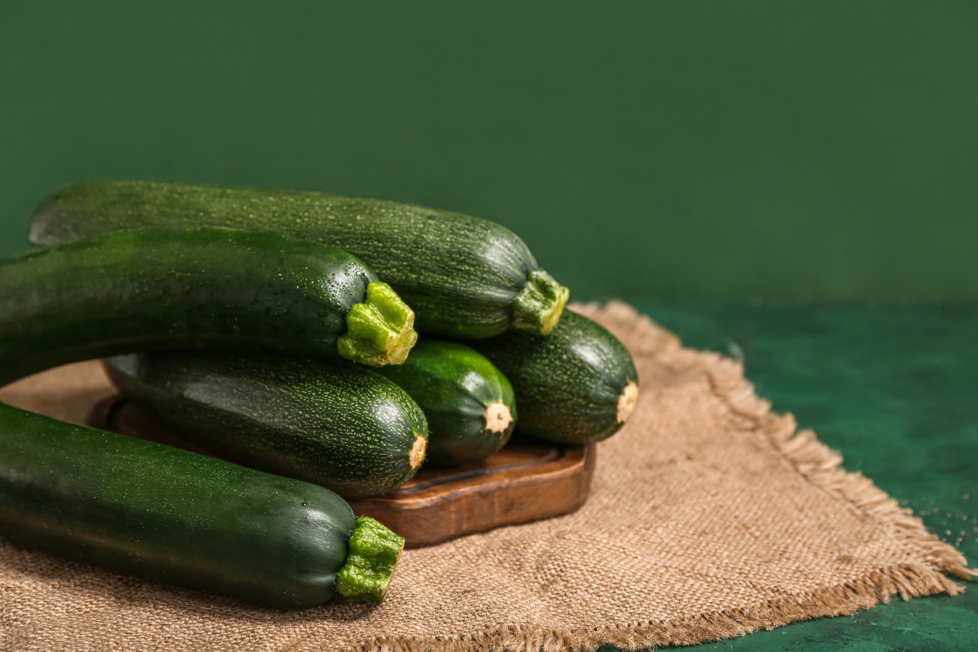 Wooden board with many fresh zucchini on green background�