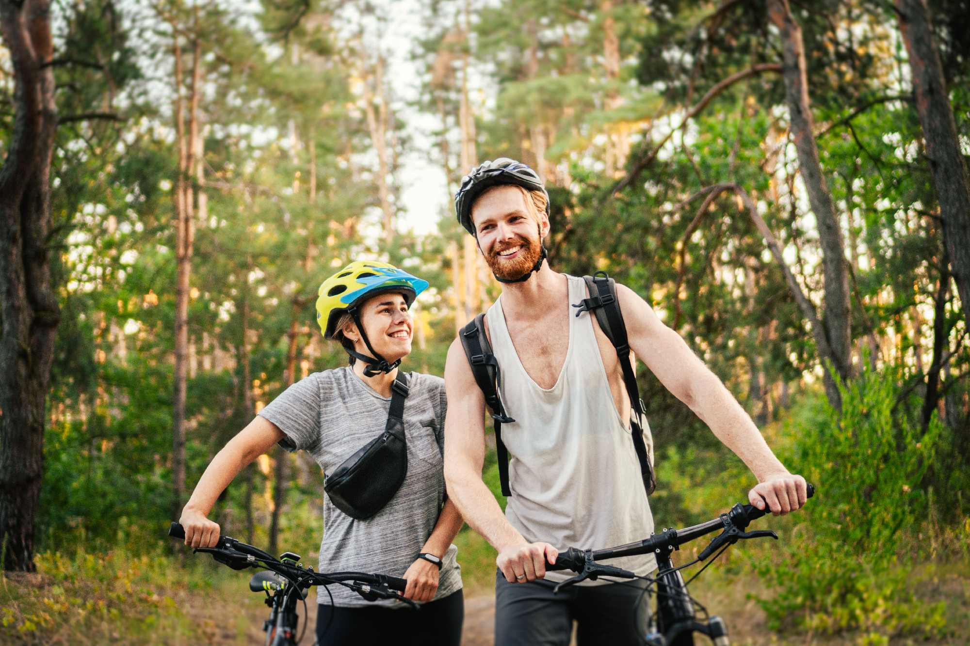 Cheerful couple pushing bicycles and walking along the forest road. Happy couple with bicycle walking through park and talking and laugh. Caucasian young man and woman riding bicycles in woods