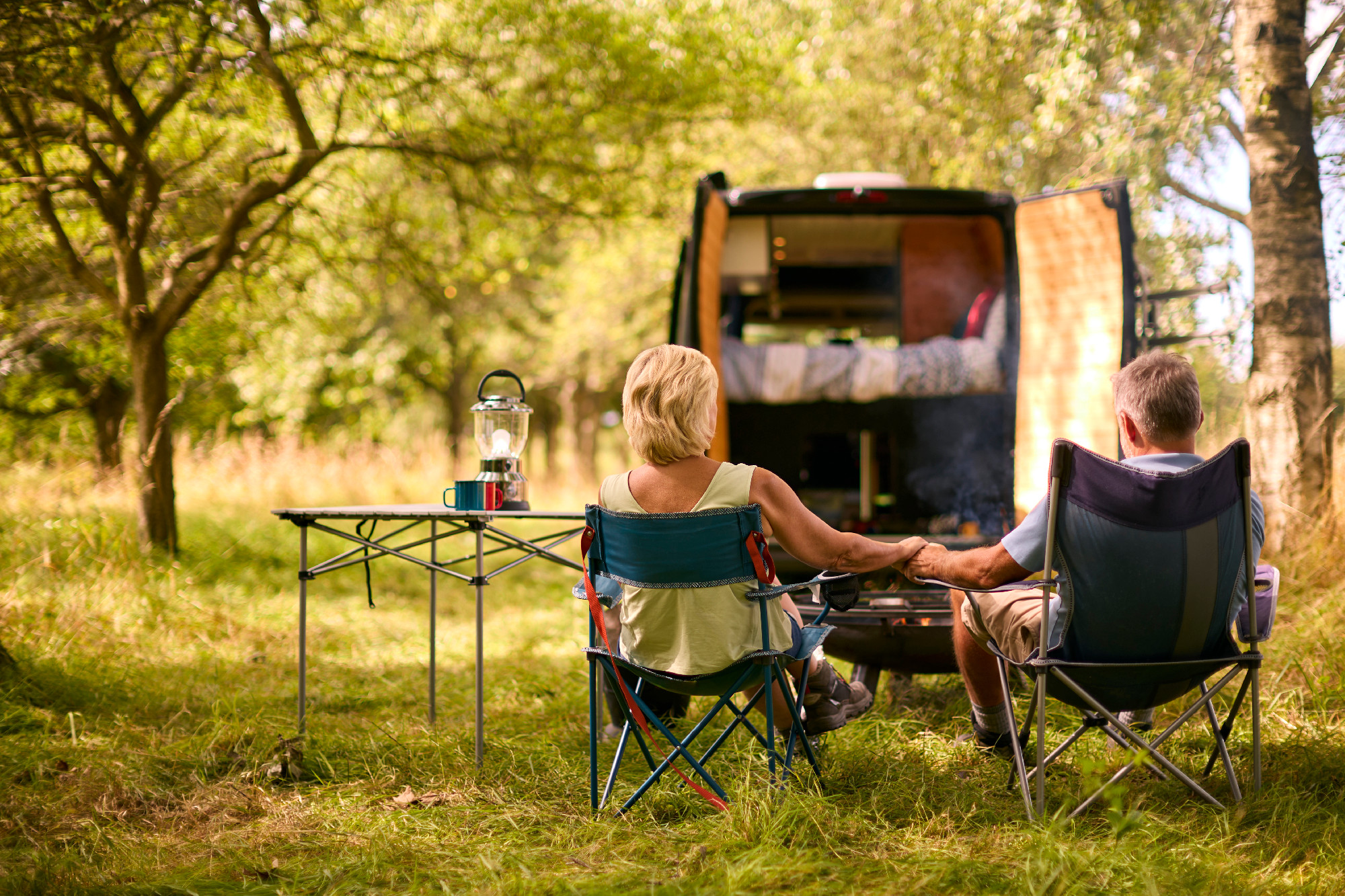 Rear View Of Loving Senior Couple Camping In Countryside With RV Holding Hands By Outdoor Fire