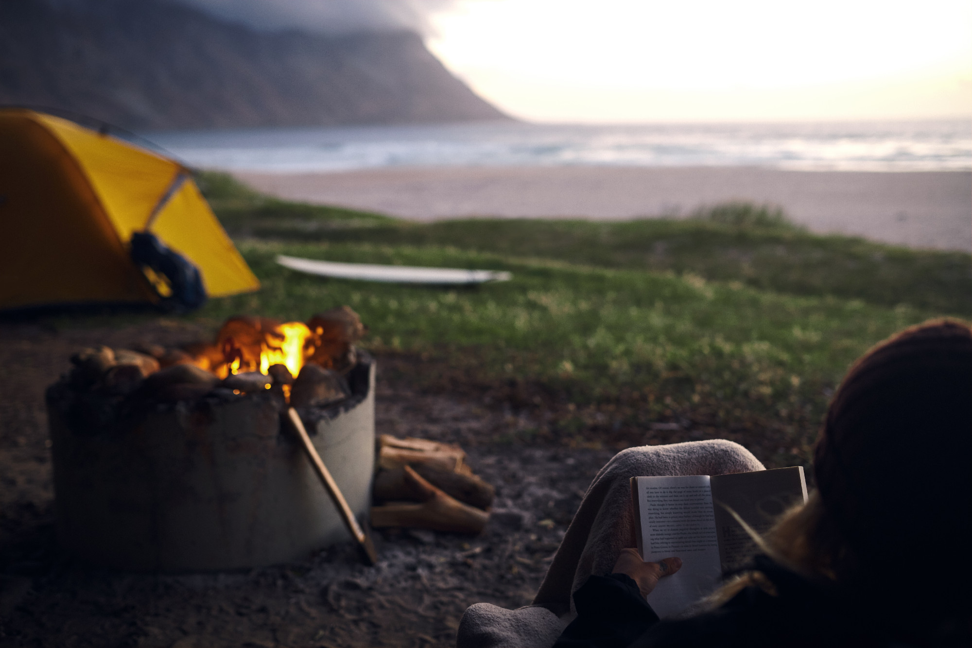 Reading, outdoor and person with book, campsite and back of college student, relax or warm in morning. Literature, fire and peace of environment, tent and camper with knowledge or learning with novel