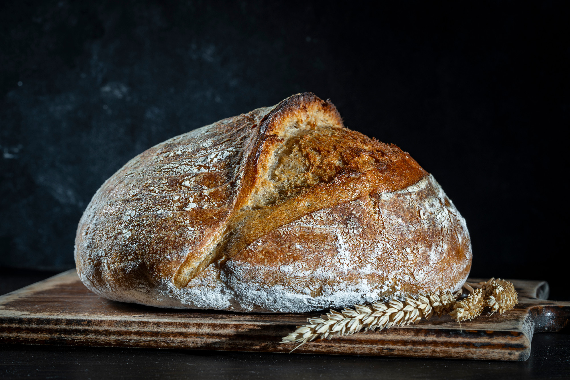Round loaf of freshly baked sourdough bread with ears of wheat on wooden cutting board, closeup. Rustic sourdough bread. Cooking healthy sourdough bread at home