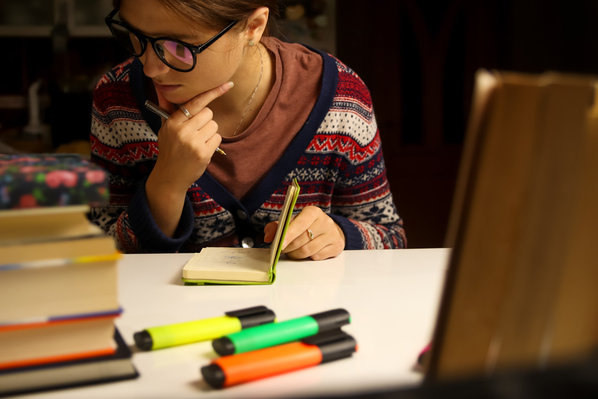 Young woman in glasses study at night, thinks. Millennial student holding light green notebook. Blurred books and markers on foreground. Education and learning, deadline concept, background