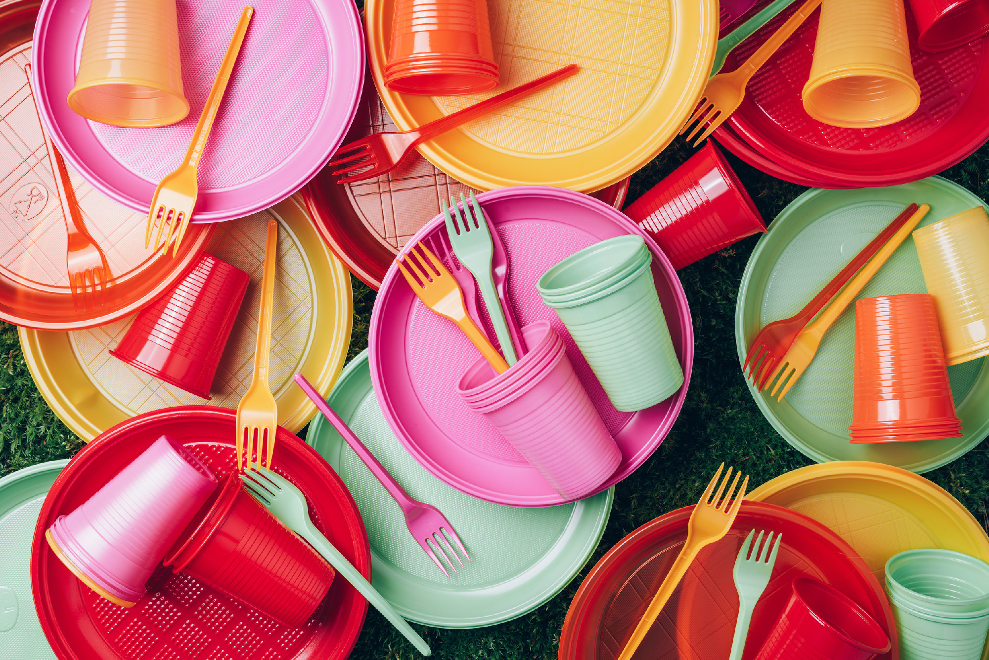 Disposable plastic tableware - plates, forks, spoons. Colored plastic disposable tableware on green grass moss background. Top view. Copy space. Birthday picnic utensil. Recycling plastic and ecology
