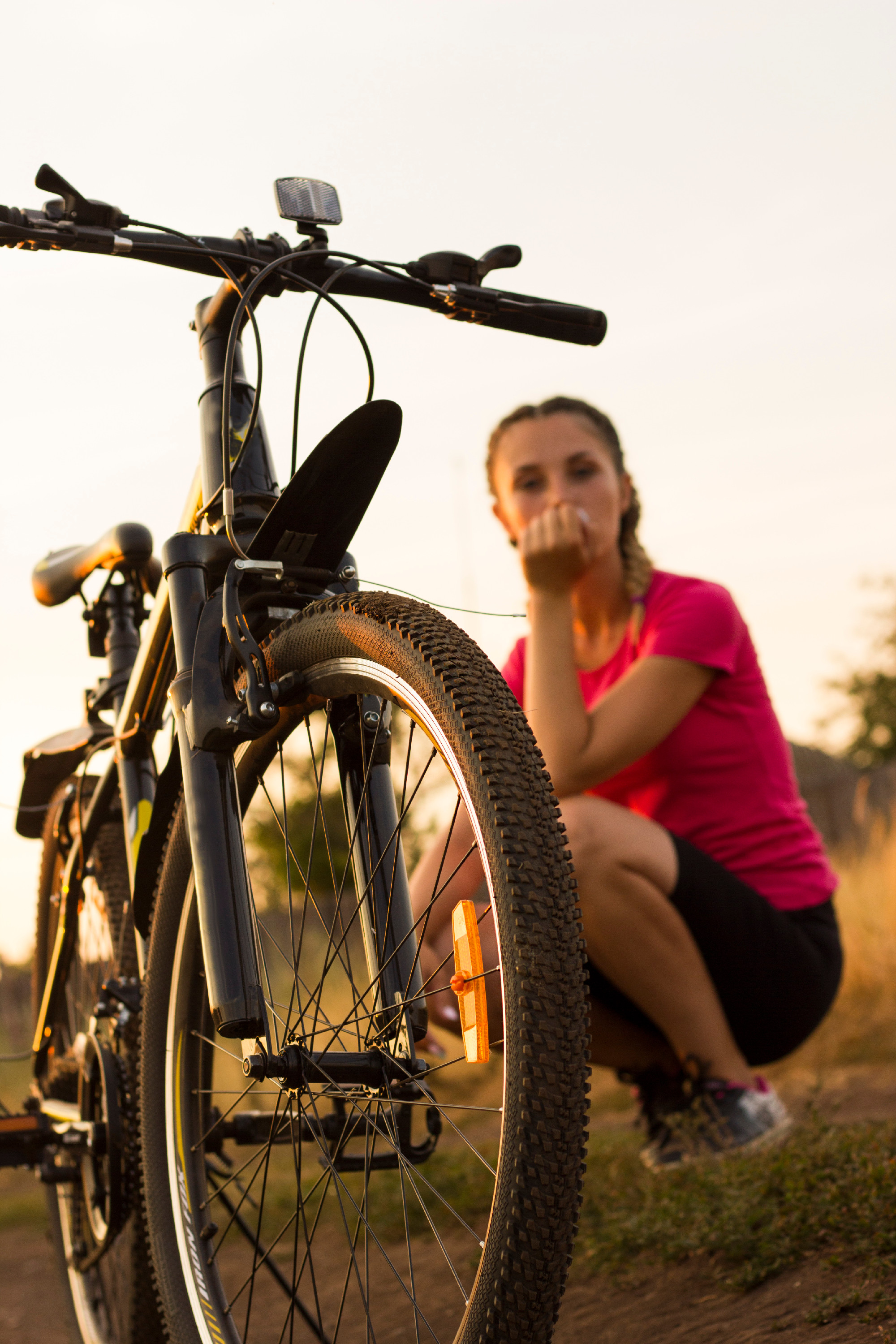 a big bicycle wheel in the foreground, a young sports girl sits in a field behind a bicycle in the background, a bicycle ride in a yellow field, the setting sun, a black speed bike