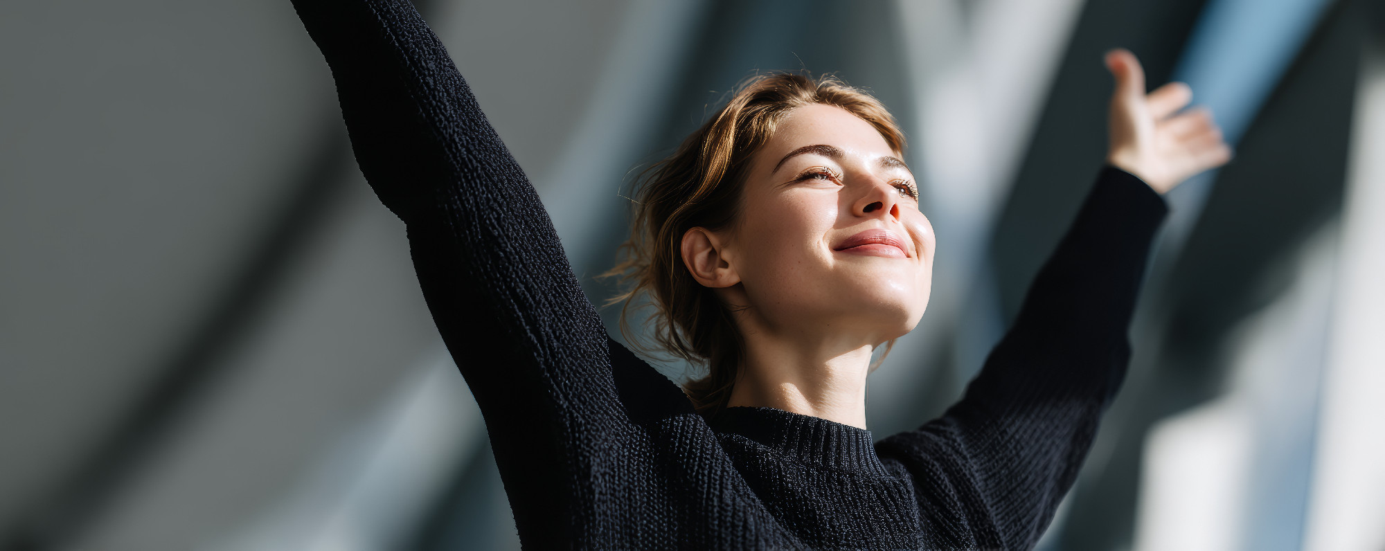 Young woman feeling joyful with arms raised, enjoying sunlight and fresh air in modern bright space