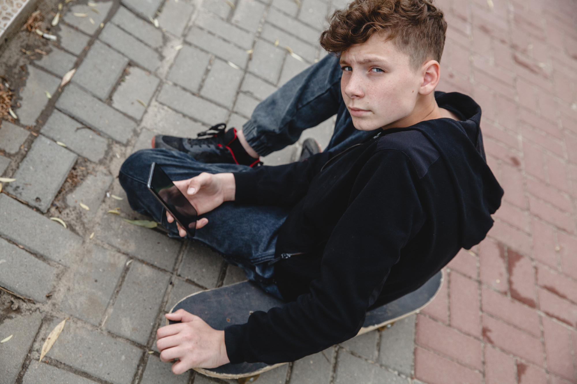 A curly-haired teen of European appearance in black hoodie sitting on an alley on a skateboard and talking on the phone