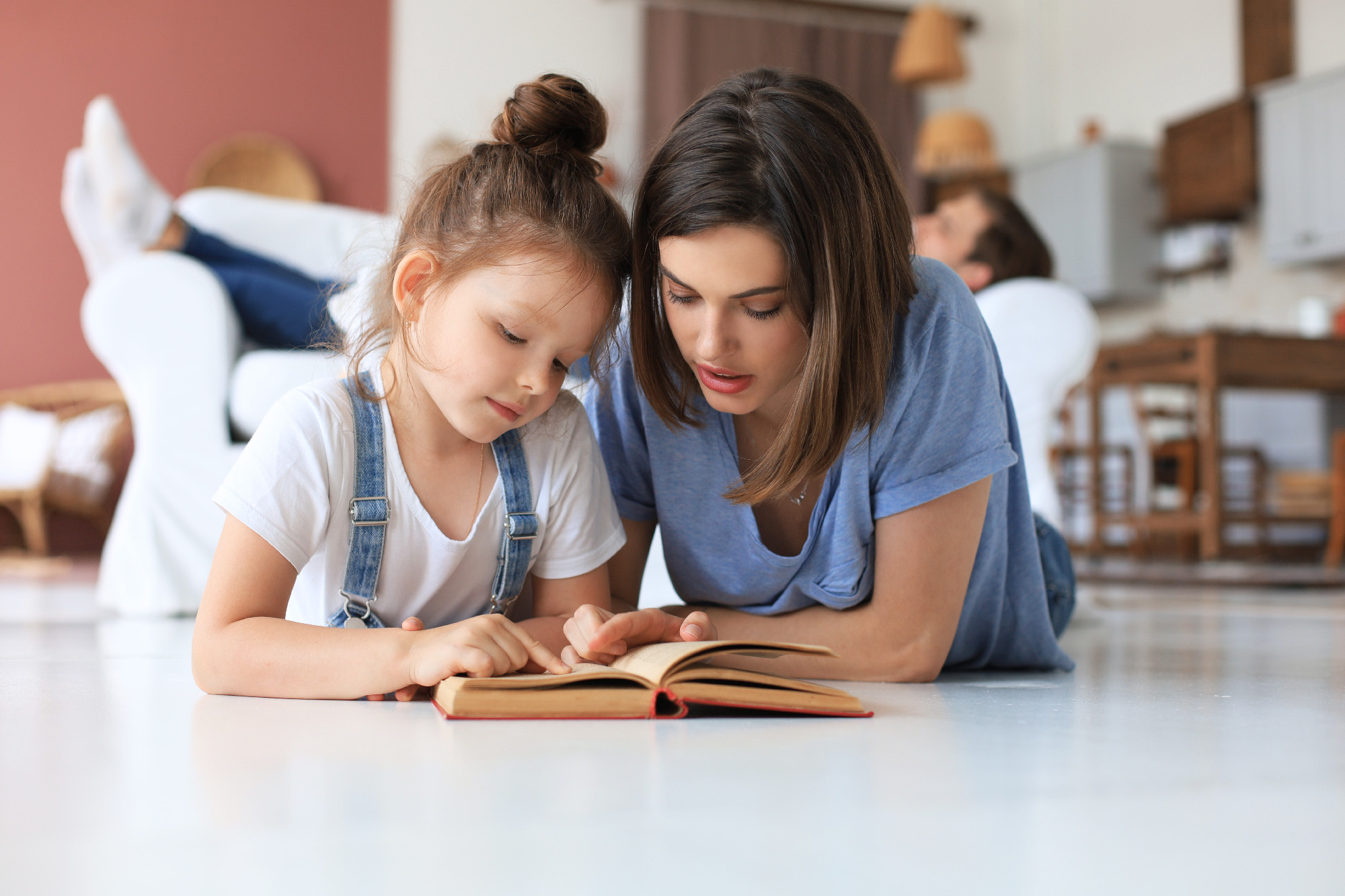 Mother and little daughter read a book together, lying on the floor in the living room.