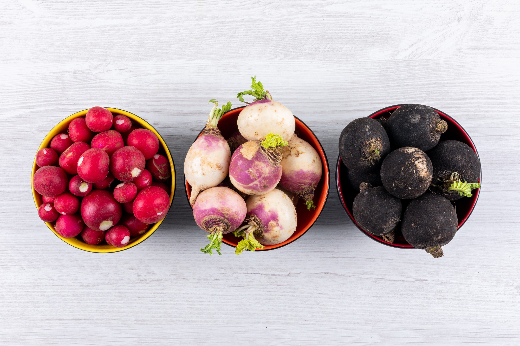 Top view fresh different color radishes in three black bowls on white wooden background. horizontal copy space for text