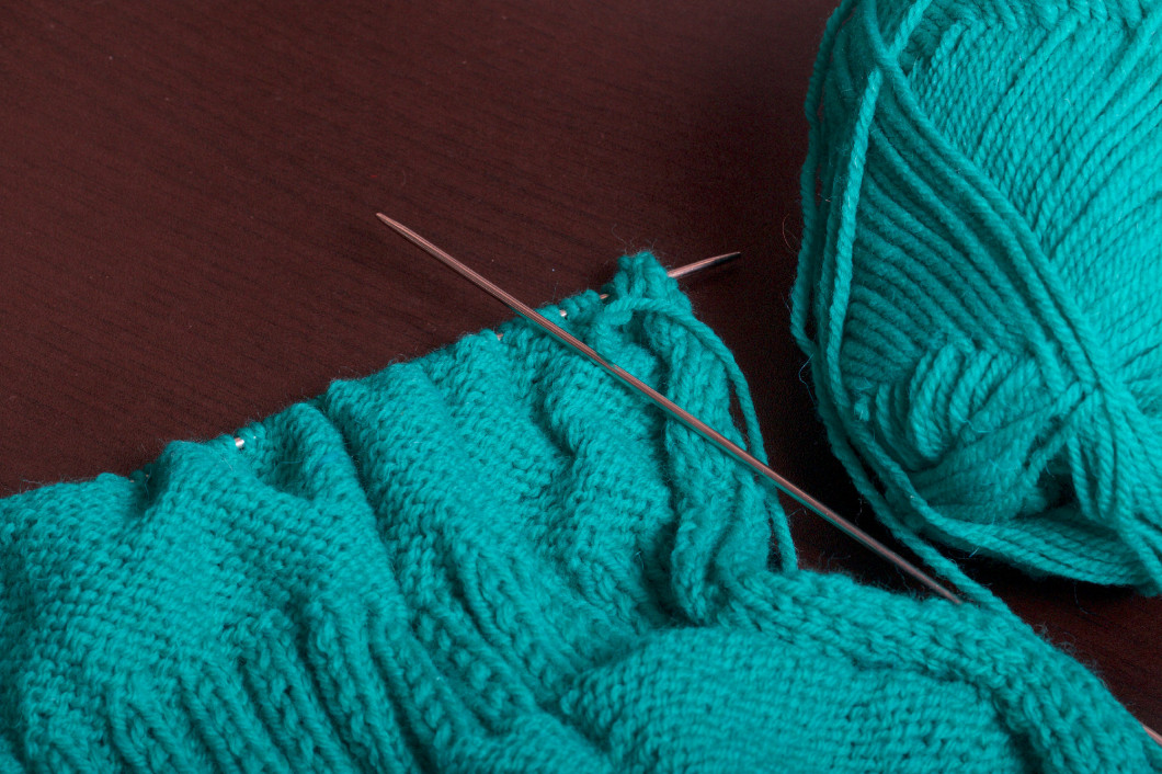 Knitting with knitting needles. On the dark surface of the table lies an unfinished knitted product with knitting needles and a ball of emerald-colored threads.