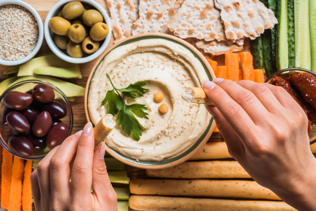 partial view of woman and arranged hummus in bowl, pita bread, cut vegetables, dried tomatoes and olives