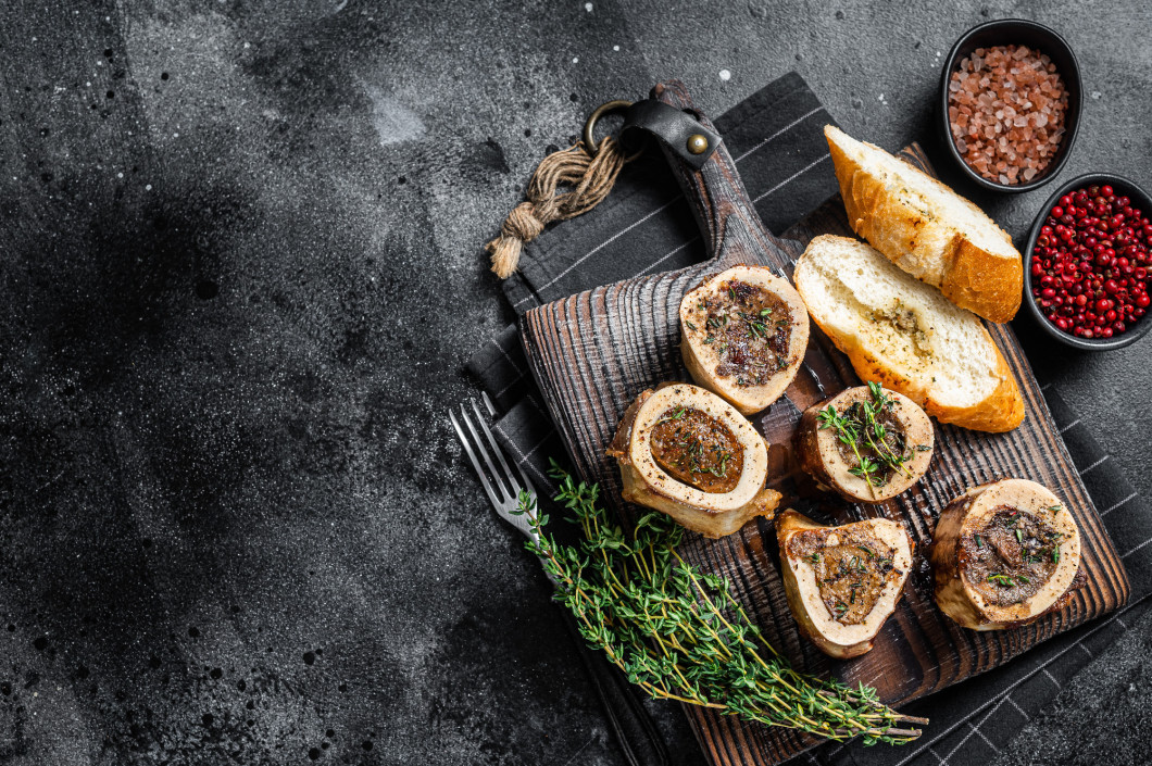 Roast marrow calf bones on wooden board with bread and herbs. Black background. Top view. Copy space