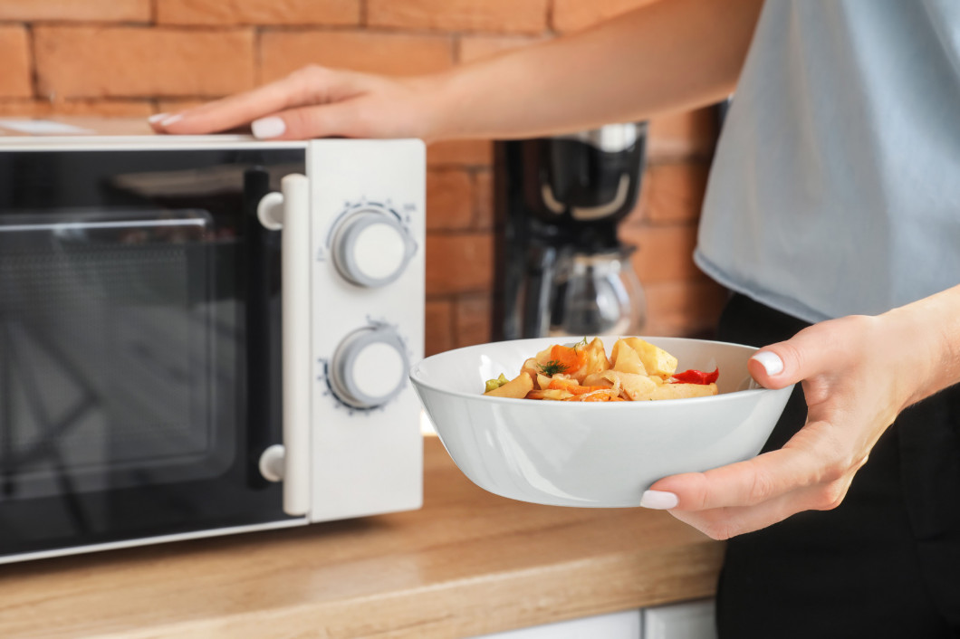 Woman holding bowl with delicious food near counter with microwave oven�