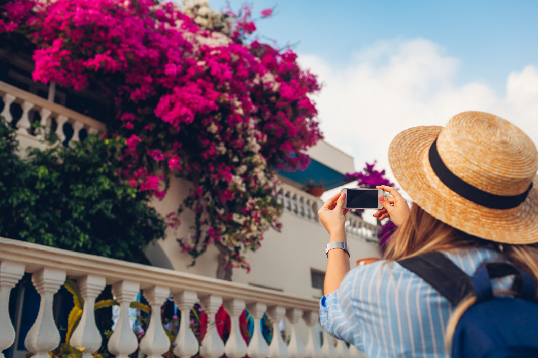 Woman traveler takes pictures of blooming bougainvillea flowers on street in Greece. Tourist enjoys landscape